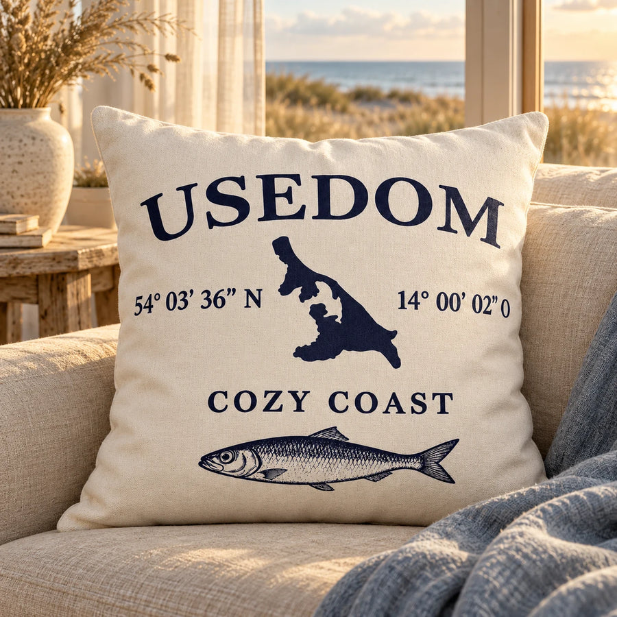 Cozy Coast Kissenhülle USEDOM 50 x 50 cm in Natur mit dunkelblauer Inselsilhouette, Koordinaten und Fischmotiv auf einem hellen Sofa mit Blick auf Dünen und Ostsee