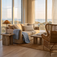 Nahaufnahme Sommerhaus-Look: Sofa mit Kissen "Riviera Beach" von Riviera Maison, hellblaue Isabel-Tagesdecke von Lene Bjerre, Rattan-Lampe von J-Line, Rattan-Sessel und Holztisch vor Dünenlandschaft
